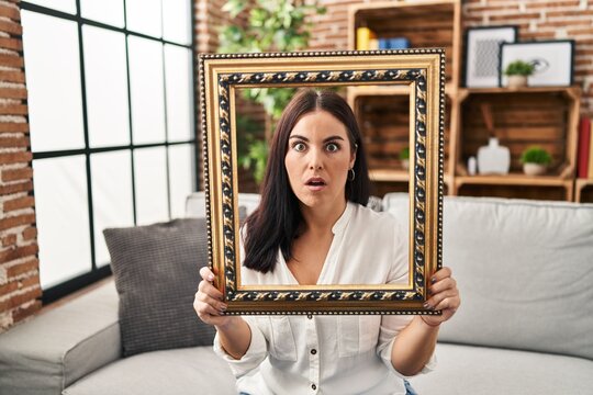 Young hispanic woman holding empty frame afraid and shocked with surprise and amazed expression, fear and excited face.