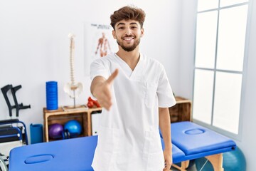 Young arab man working at pain recovery clinic smiling friendly offering handshake as greeting and welcoming. successful business.