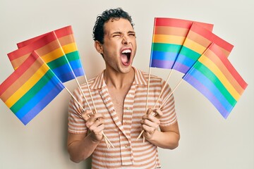 Handsome man wearing make up holding rainbow lgbtq flags angry and mad screaming frustrated and...