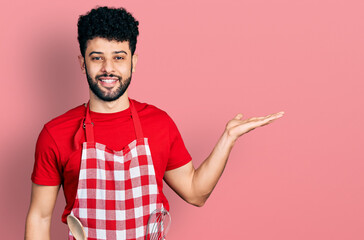 Young arab man with beard wearing cook apron smiling cheerful presenting and pointing with palm of hand looking at the camera.