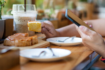 Close up to slices of toast bread and butter with steamed Thai tea custard topping in hand of woman using fork to eat for breakfast during using mobile phone. Relaxing with food and technology concept