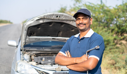 Portriat shot smiling car mechanic stading in front of borken on roadside by looking at camera -...