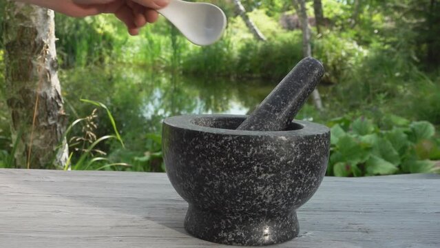 Hand is putting salt and spices into grey stone mortar in slow motion outdoors