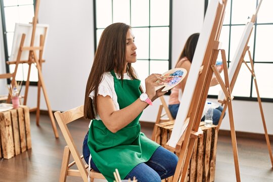 Young hispanic artist women painting on canvas at art studio looking to side, relax profile pose with natural face and confident smile.