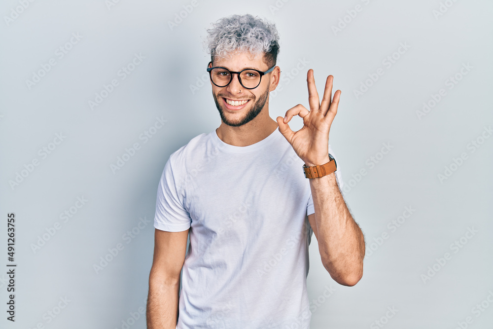 Wall mural Young hispanic man with modern dyed hair wearing white t shirt and glasses smiling positive doing ok sign with hand and fingers. successful expression.