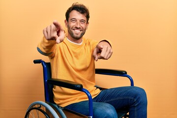 Handsome man with beard sitting on wheelchair pointing to you and the camera with fingers, smiling...