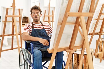 Young handsome man with beard at art studio sitting on wheelchair scared and amazed with open mouth for surprise, disbelief face