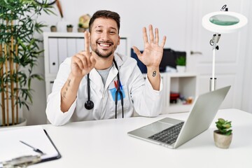 Young doctor working at the clinic using computer laptop showing and pointing up with fingers number six while smiling confident and happy.