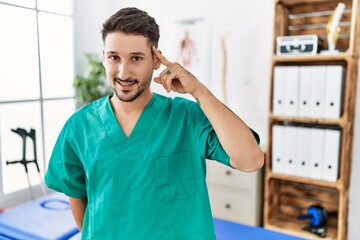 Young physiotherapist man working at pain recovery clinic smiling pointing to head with one finger, great idea or thought, good memory