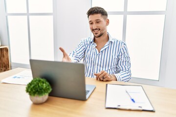 Young handsome man with beard working at the office using computer laptop smiling friendly offering handshake as greeting and welcoming. successful business.