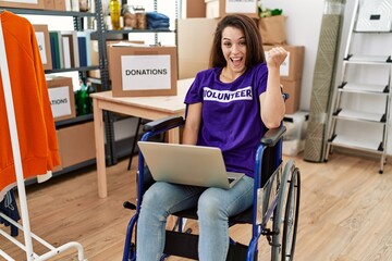 Young brunette woman as volunteer on donations stand sitting on wheelchair screaming proud, celebrating victory and success very excited with raised arms