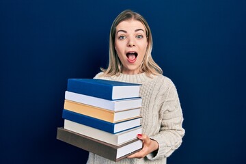 Beautiful caucasian woman holding a pile of books celebrating crazy and amazed for success with open eyes screaming excited.