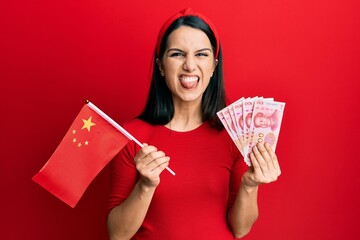 Young hispanic woman holding china flag and yuan banknotes sticking tongue out happy with funny...