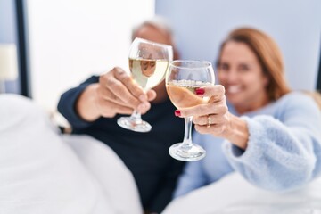 Middle age man and woman couple drinking wine lying on bed at bedroom