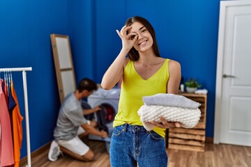 Young brunette woman holding folded laundry smiling happy doing ok sign with hand on eye looking through fingers