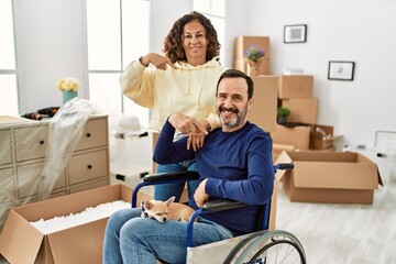 Middle age hispanic couple and dog sitting on wheelchair at new home pointing finger to one self...