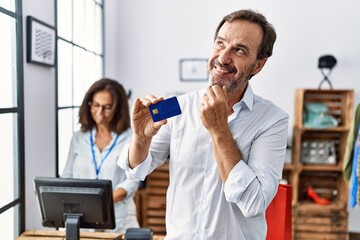Hispanic man holding credit card at retail shop serious face thinking about question with hand on chin, thoughtful about confusing idea