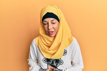 Middle age hispanic woman wearing traditional islamic hijab scarf with hand on stomach because indigestion, painful illness feeling unwell. ache concept.