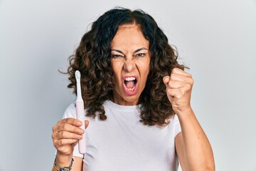 Middle age hispanic woman holding electric toothbrush annoyed and frustrated shouting with anger, yelling crazy with anger and hand raised