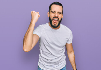 Young man with beard wearing casual white t shirt angry and mad raising fist frustrated and furious...