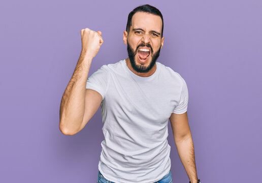 Young man with beard wearing casual white t shirt angry and mad raising fist frustrated and furious while shouting with anger. rage and aggressive concept.