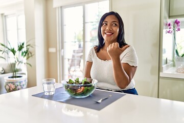 Young hispanic woman eating healthy salad at home smiling with happy face looking and pointing to the side with thumb up.