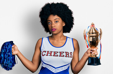 Young african american woman wearing cheerleader uniform holding pompom and trophy relaxed with serious expression on face. simple and natural looking at the camera.