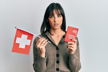 Young hispanic woman holding swiss flag and passport relaxed with serious expression on face. simple and natural looking at the camera.