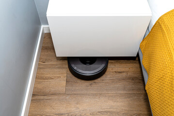 A modern robotic vacuum cleaner cleans vinyl panels under bedroom furniture, an autonomous cleaning...