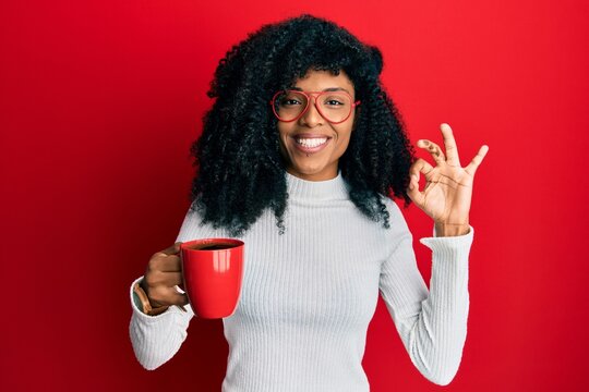 African american woman with afro hair holding coffee doing ok sign with fingers, smiling friendly gesturing excellent symbol