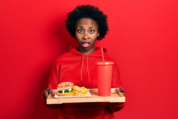 Young african american woman eating a tasty classic burger with fries and soda in shock face, looking skeptical and sarcastic, surprised with open mouth