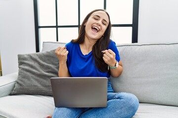 Naklejka premium Young brunette woman using laptop at home very happy and excited doing winner gesture with arms raised, smiling and screaming for success. celebration concept.