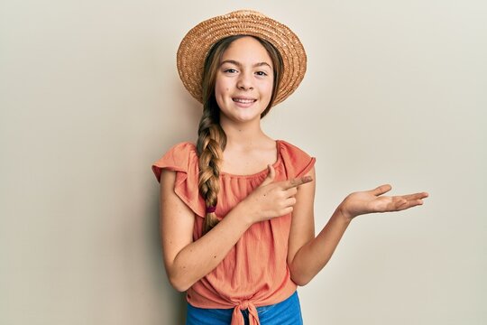 Beautiful brunette little girl wearing summer hat amazed and smiling to the camera while presenting with hand and pointing with finger.