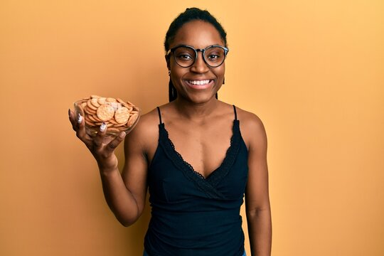African american woman with braided hair holding salty biscuits bowl looking positive and happy standing and smiling with a confident smile showing teeth
