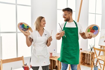 Young caucasian couple smiling happy holding paintbrush and palette at art studio.