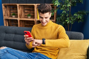 Young hispanic man using smartphone and looking watch at home