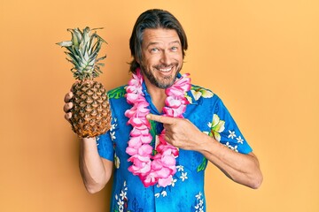 Middle age handsome man wearing hawaiian lei holding pineapple smiling happy pointing with hand and...