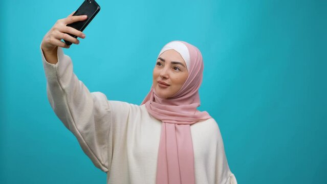 Young Muslim woman in hijab taking selfie. Isolated studio shot on blue background