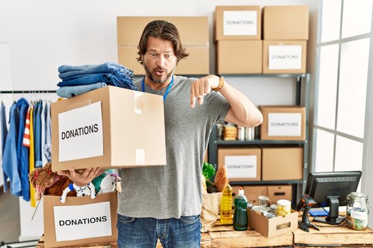 Handsome middle age man holding donations box for charity at volunteer stand pointing down with fingers showing advertisement, surprised face and open mouth