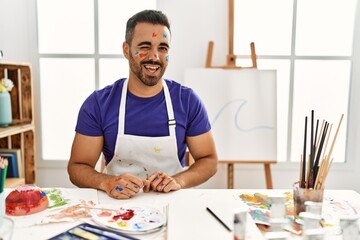 Young hispanic man with beard at art studio with painted face winking looking at the camera with sexy expression, cheerful and happy face.