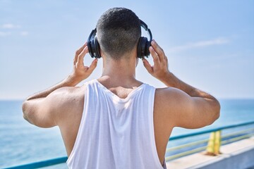 Hispanic sports man wearing headphones listening to music by the sea on a sunny day