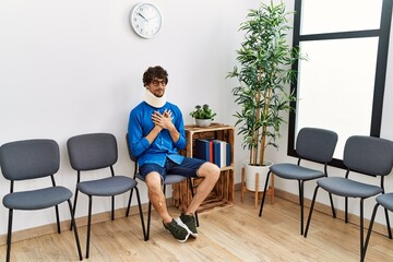 Young hispanic man sitting at doctor waiting room with neck injury smiling with hands on chest with closed eyes and grateful gesture on face. health concept.