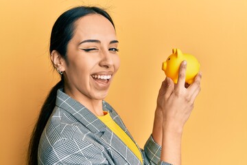 Young hispanic girl looking to piggy bank winking looking at the camera with sexy expression,...