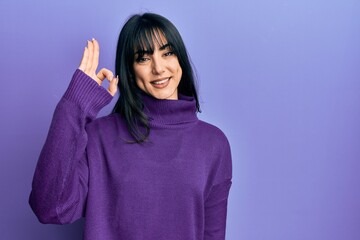 Fototapeta premium Young brunette woman with bangs wearing turtleneck sweater smiling positive doing ok sign with hand and fingers. successful expression.