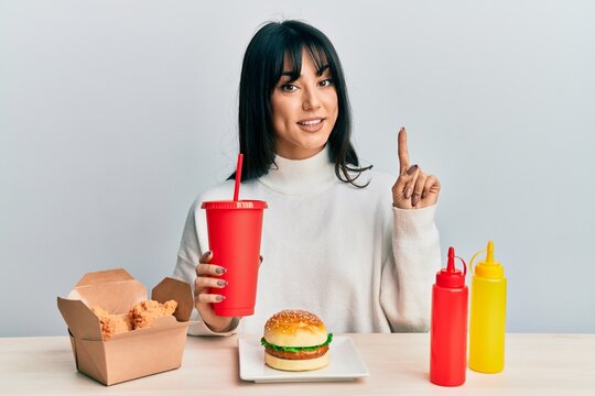Young brunette woman with bangs eating a tasty classic burger with ketchup and mustard smiling with an idea or question pointing finger with happy face, number one