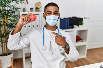 Fototapeta na wymiar Young indian doctor holding virus toy wearing safety mask smiling happy pointing with hand and finger