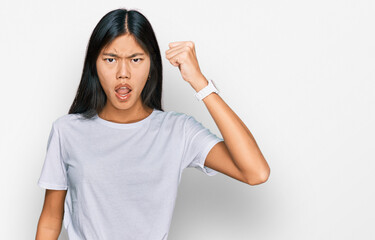 Beautiful young asian woman wearing casual white t shirt angry and mad raising fist frustrated and furious while shouting with anger. rage and aggressive concept.