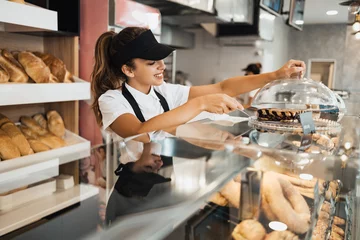 Gordijnen Mooie jonge en gelukkige vrouwelijke werknemer die werkt in een moderne bakkerij of fastfoodrestaurant. © Dusko