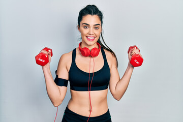 Young hispanic girl wearing sportswear using dumbbells winking looking at the camera with sexy...