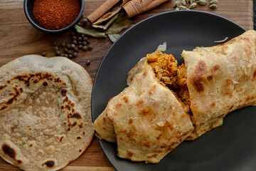Chicken salomie or roti on a black plate with curry powder, cinnamon sticks, green cardamom in background